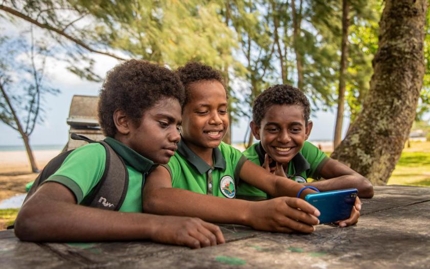 Three boys are smiling and gathered around together looking at a phone screen held in the hands of the boy in the middle of the group.